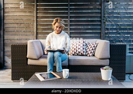 Donna sorridente che scrive nel diario mentre si siede sul divano a. giardino sul tetto Foto Stock