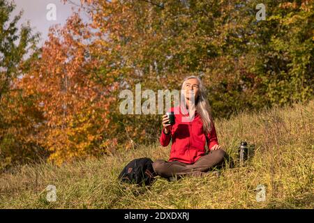 Donna sorridente con coppa thermos seduta in montagna alle colline alpine, Germania Foto Stock