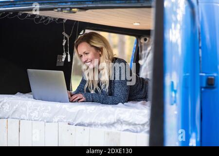 Donna sorridente che usa il computer portatile mentre si sdraiava sul letto in pulmino Foto Stock