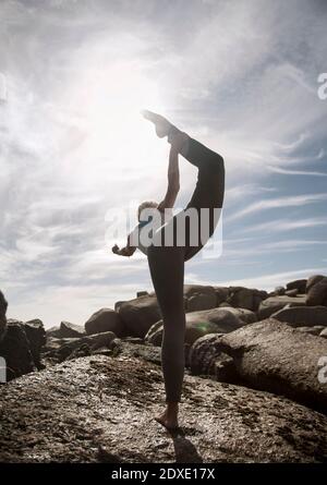Donna flessibile che pratica Natarajasana sulla formazione rocciosa in spiaggia Foto Stock