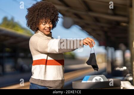 Giovane donna gettando la maschera protettiva usata nel cestino della spazzatura alla stazione Foto Stock