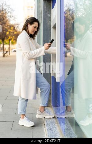 Elegante imprenditore femminile che utilizza lo smartphone mentre si ritirano i soldi da BANCOMAT Foto Stock