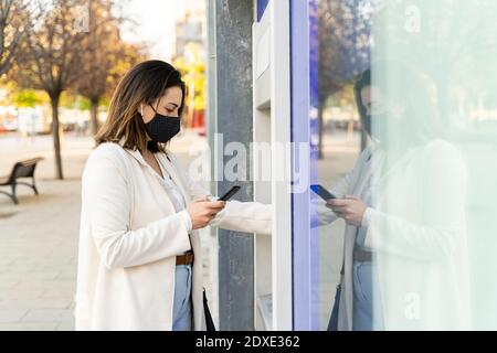 Imprenditrice femminile che utilizza lo smartphone mentre ritira denaro dagli ATM in città durante la pandemia Foto Stock