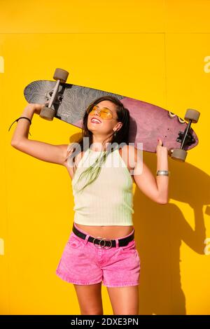 Ritratto di giovane donna in piedi di fronte al muro giallo con longboard in mani Foto Stock