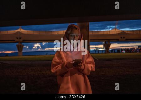 Giovane donna con capelli rosa che indossa una camicia con cappuccio rosa che regge smartphone in città di notte Foto Stock