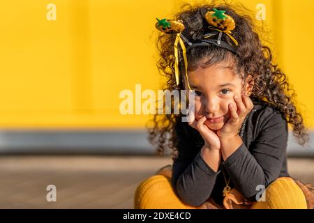 Ragazza carina che indossa la fascia di Halloween seduta con la testa nelle mani contro parete gialla Foto Stock