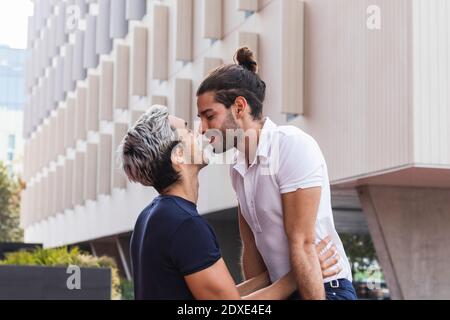 Uomo sorridente che guarda il compagno gay mentre bacia contro la costruzione in città Foto Stock