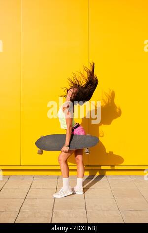 Giovane donna che lancia i capelli davanti alla parete gialla con longboard in mano Foto Stock