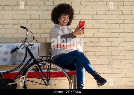 Donna felice che prende selfie mentre si siede sulla panchina in bicicletta contro muro di mattoni Foto Stock