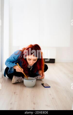 Donna d'affari redhead che mangia noodle mentre si siede sul pavimento in ufficio Foto Stock