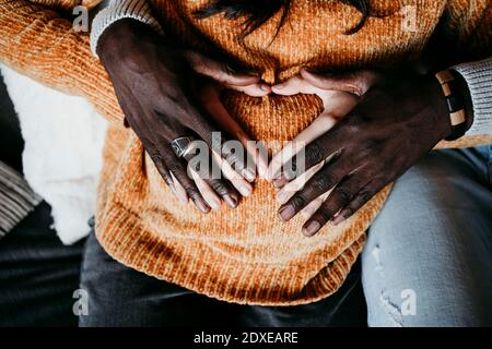 L'uomo e la donna incinta formano la forma del cuore con le mani sopra pancia a casa Foto Stock