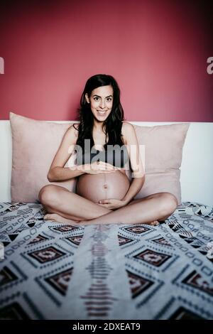 Donna incinta sorridente seduta sul letto contro il muro rosso a. casa Foto Stock