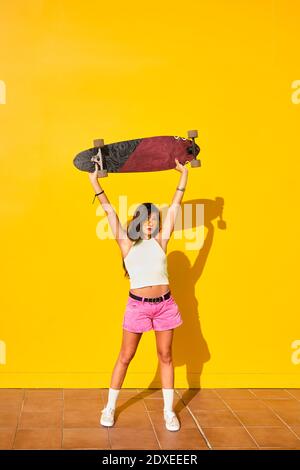 Ritratto di giovane donna in piedi di fronte al muro giallo con longboard in mani Foto Stock