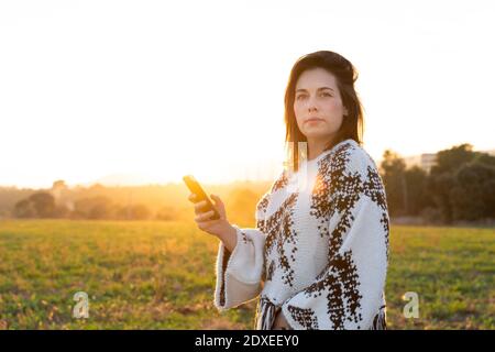 Bella giovane donna che tiene il telefono cellulare mentre si guarda via durante tramonto Foto Stock