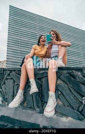 Sorridere le amiche utilizzando il telefono cellulare mentre si siede contro la parete Foto Stock