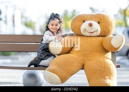 Ragazza sorridente seduta con orsacchiotto sulla panchina Foto Stock