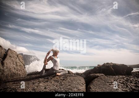 Donna bionda flessibile che pratica yoga sulla formazione rocciosa in spiaggia Foto Stock