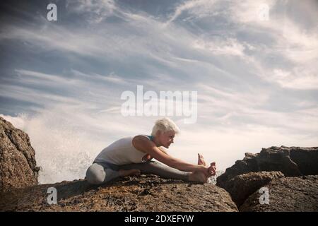 Donna bionda flessibile che pratica yoga sulla formazione rocciosa in spiaggia contro il cielo Foto Stock