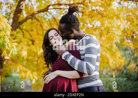 Uomo africano baciando donna incinta felice nel parco durante l'autunno Foto Stock