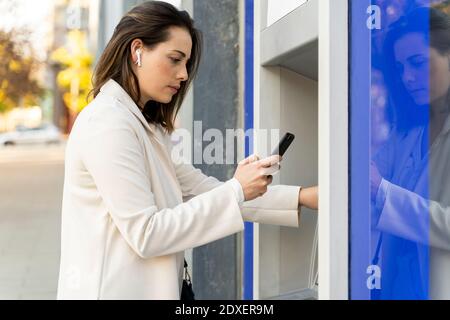 Donna d'affari che usa lo smartphone mentre ritira i soldi dal ATM in città Foto Stock