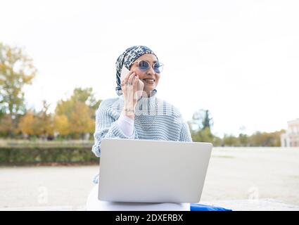 Donna sorridente che indossa un copricapo che parla sul telefono cellulare mentre è seduto all'aperto Foto Stock