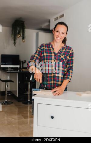 Donna sorridente che usa un trapano elettrico su materiale di legno mentre si è in piedi da cabinet a casa Foto Stock