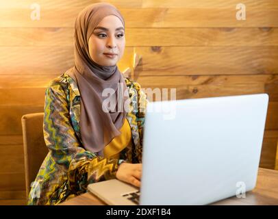 Giovane donna che indossa hijab usando il computer portatile mentre si siede al bar Foto Stock
