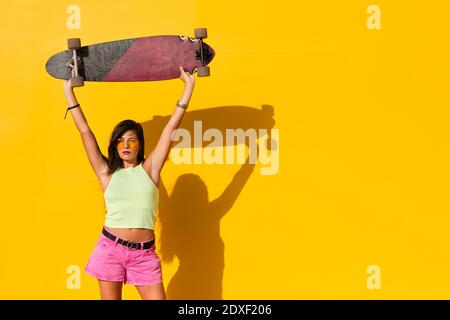 Ritratto di giovane donna in piedi di fronte al muro giallo con longboard in mani Foto Stock