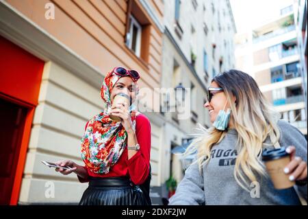 Sorridendo amici femminili con tazze usa e getta che camminano in città durante COVID-19 Foto Stock