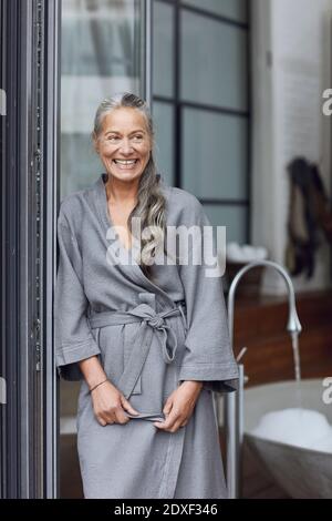 Donna matura sorridente che indossa accappatoio appoggiato da una porta in vetro bagno Foto Stock