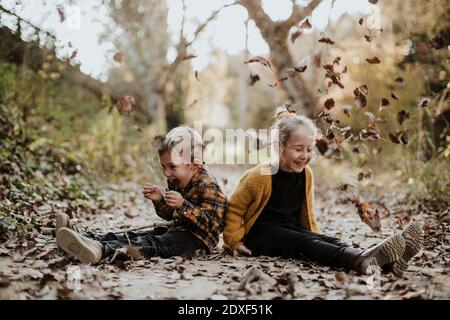 Ragazza allegra e ragazzo che gioca con foglie secche caduto mentre seduto sul sentiero nella foresta Foto Stock