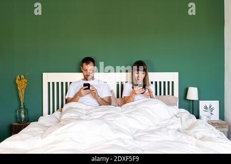 Coppia che utilizza il telefono cellulare mentre si è seduti a letto a. casa Foto Stock