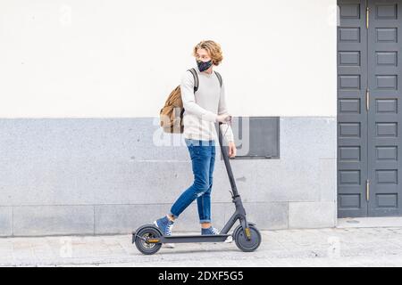 Giovane uomo che indossa una maschera protettiva in piedi con spinta elettrica scooter sul sentiero Foto Stock