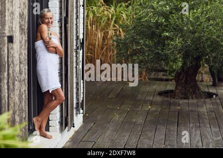 Donna matura felice che indossa un asciugamano appoggiato sulla porta della schiena iarda Foto Stock