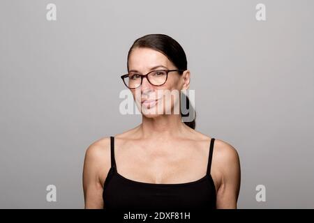 Donna matura sorridente in cima nera con occhiali contro il grigio sfondo Foto Stock