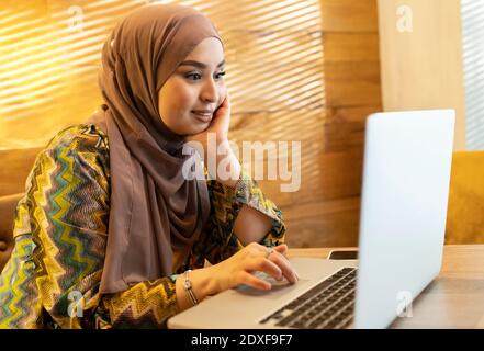 Giovane donna che indossa hijab seduto con la mano sul mento mentre lavorare su un computer portatile al bar Foto Stock