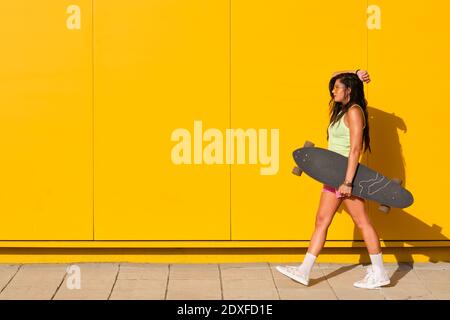 Giovane donna che cammina lungo il muro giallo con la tavola lunga in mano Foto Stock