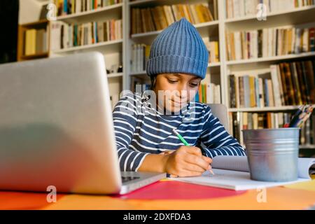 Ragazzo sorridente che indossa un cappello a maglia scritto nel libro mentre si siede a casa Foto Stock