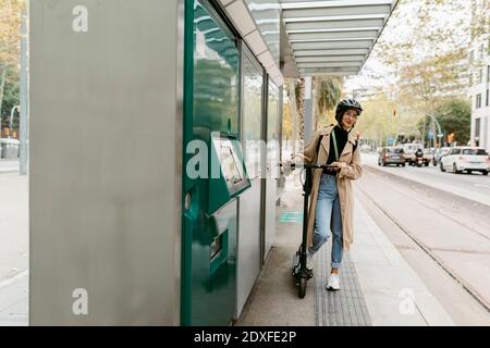 Donna che indossa casco da ciclismo in piedi con scooter elettrico push mentre attesa alla stazione del tram in città Foto Stock