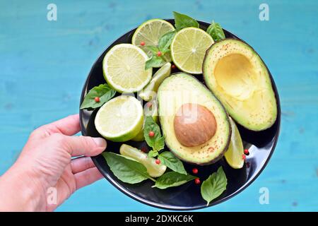 Mano bianca della donna caucasica appesa ad un piatto nero pieno di cibo sano. Avocado, lime a fette, limoni e menta su sfondo blu. Foto Stock