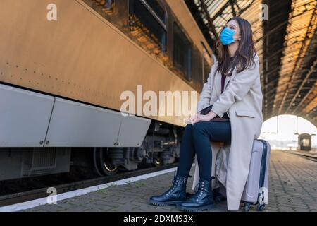 Solitaria ragazza triste in maschera medica si siede sulla valigia in attesa di treno. Viaggiatore femminile alla stazione ferroviaria vuota Foto Stock