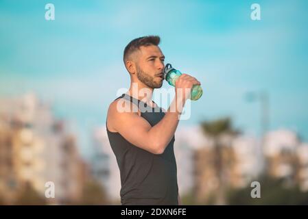 Giovane uomo in forma fisica che beve acqua da una bottiglia riutilizzabile durante un allenamento. Bere mentre si esercita. Disidratazione durante l'esercizio Foto Stock