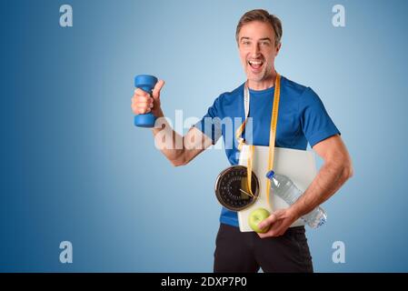 Uomo atletico con scala e nastro di misurazione appeso al colletto acqua e mela vestita di abbigliamento sportivo e gradiente isolato sfondo blu Foto Stock