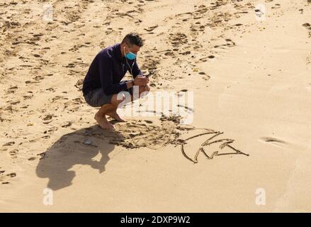 Las Palmas, Gran Canaria, Isole Canarie, Spagna. 24 dicembre 2020. Un turista che indossa una maschera faccia scatta una foto di un albero di natale disegnato nella sabbia sulla spiaggia della città a Las Palmas su Gran Canaria; una destinazione popolare di sole di inverno/Natale per i turisti britannici. Credit: Alan Dawson/Alamy Live News Foto Stock