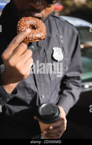 vista ritagliata del poliziotto afroamericano con la tazza di carta mangiare ciambella su sfondo sfocato all'aperto Foto Stock