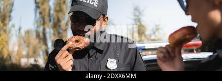 ufficiale di polizia afroamericano mangiare ciambella con collega offuscato in primo piano all'aperto, banner Foto Stock