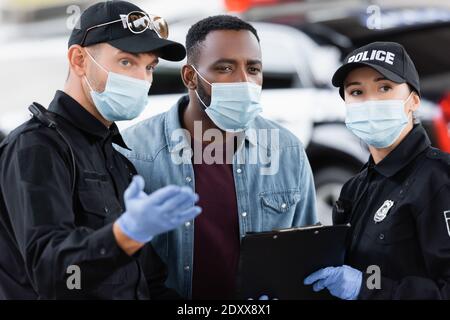 Vittima afroamericana in maschera medica in piedi vicino agenti di polizia con gli appunti sulla strada urbana Foto Stock
