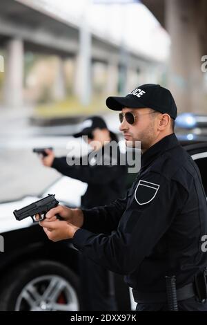 Ufficiale di polizia in occhiali da sole che tengono l'arma da fuoco vicino al collega e l'auto su sfondo sfocato all'aperto Foto Stock