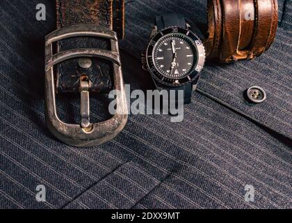 Immagine di una cintura in pelle, di un orologio nero e cromato e di un bracciale in pelle sopra un gilet a righe grigio Foto Stock