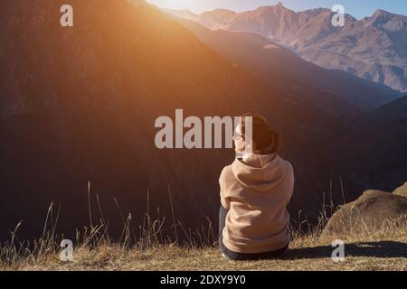 La brunetta con gli occhiali da sole si trova sul bordo marrone della collina dell'erba e guarda il paesaggio collinare sotto la luce del sole in autunno vista lato giorno Foto Stock